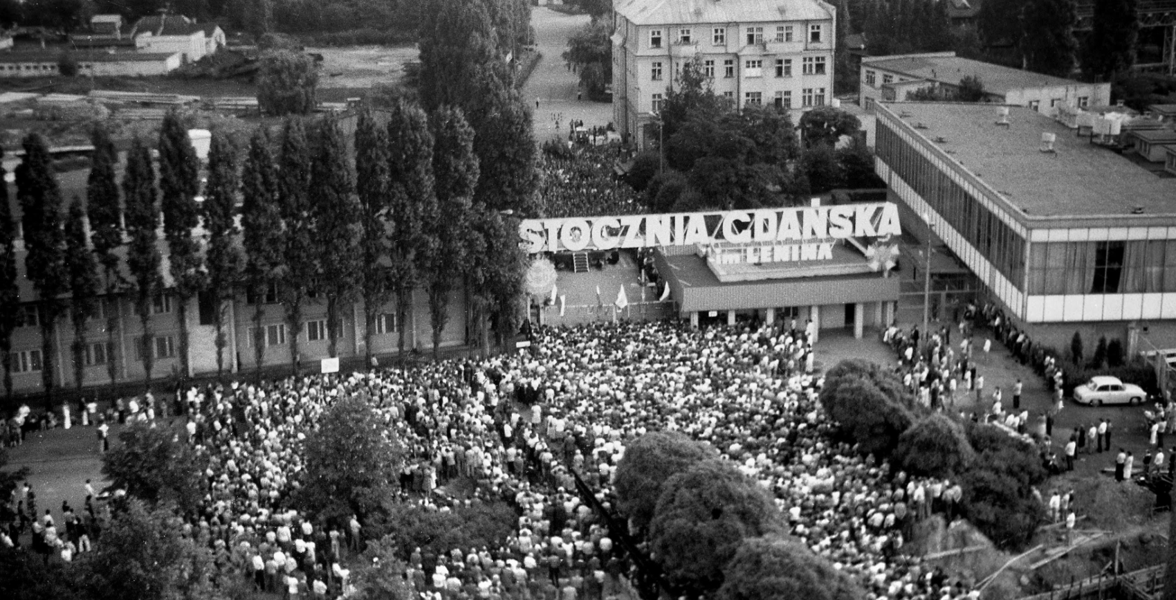 Tłum ludzi stojących pod bramą stoczni. Widoczny duży napis "Stocznia Gdańska im. Lenina"